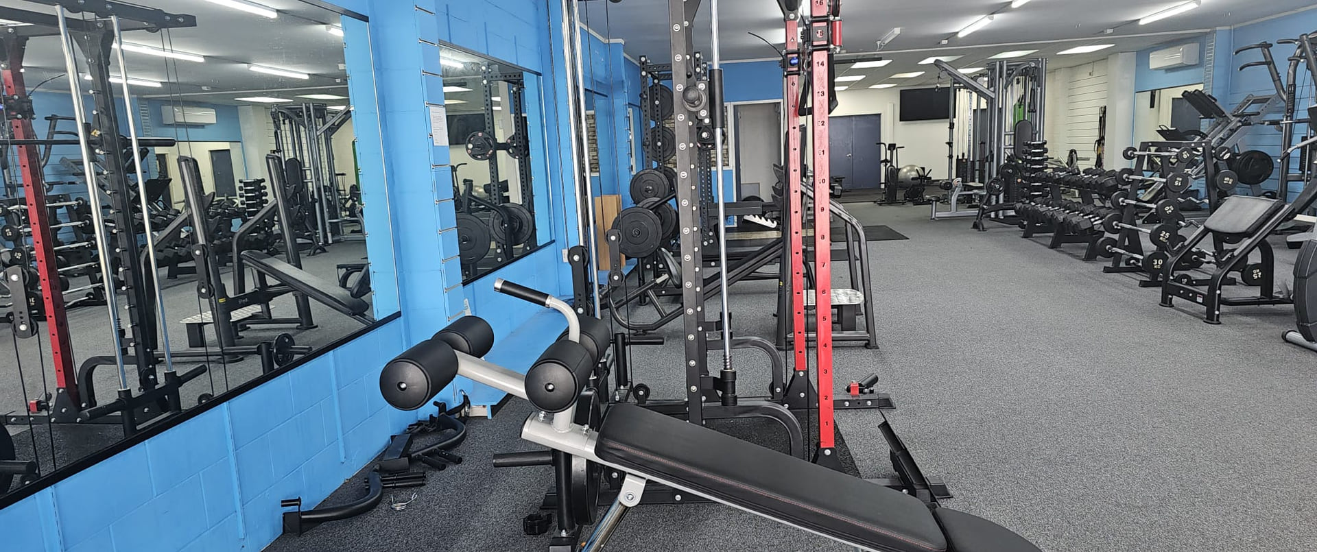 The main floor at Better Fitness Putāruru: a power rack, wall mirrors, two racks of dumbbells and a run of plate loaded machines along the blue painted brick